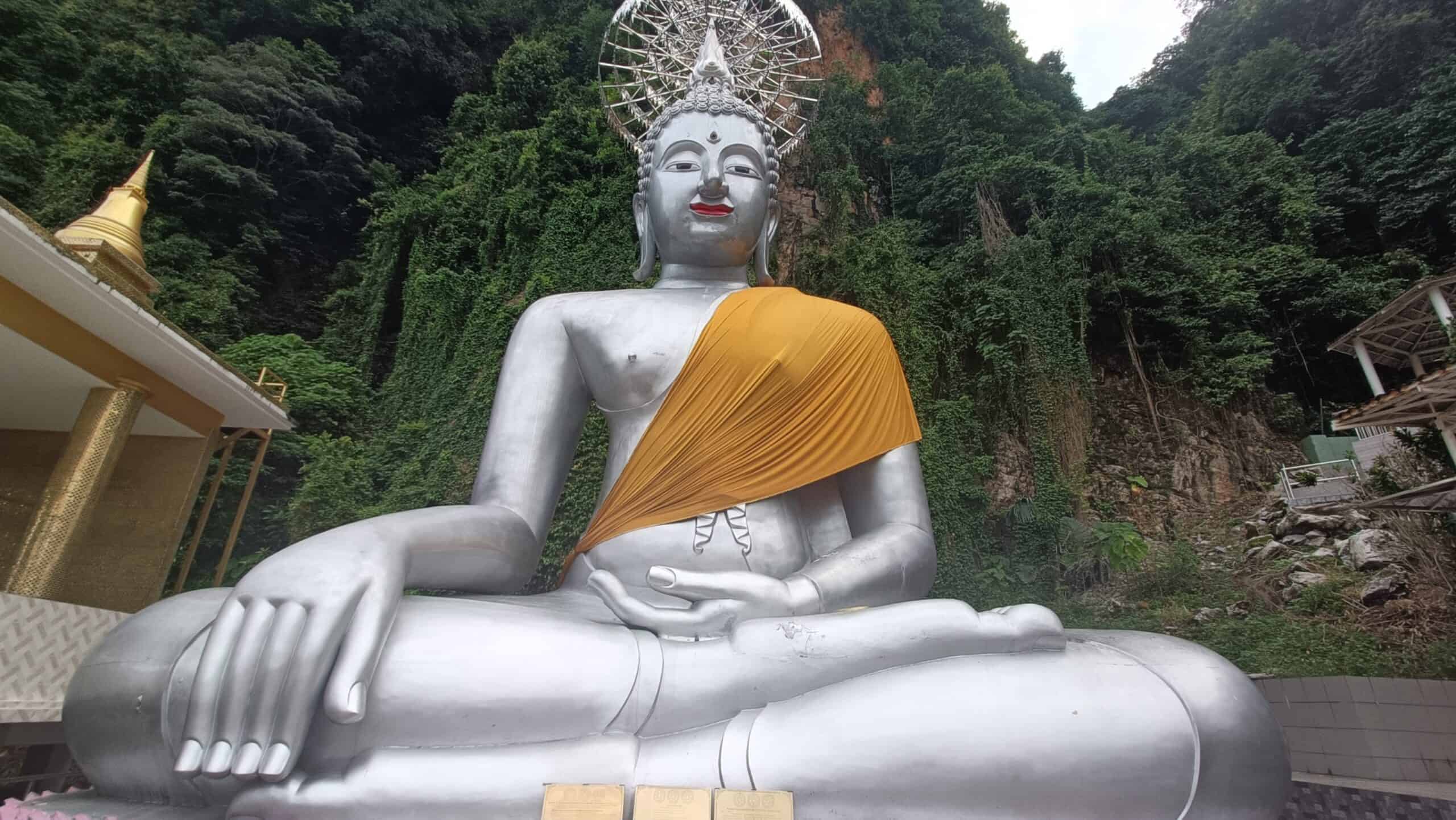 Giant silver seated Buddha statue with orange robe at Wat Puthanimittam Temple Ipoh with lush green mountains behind 