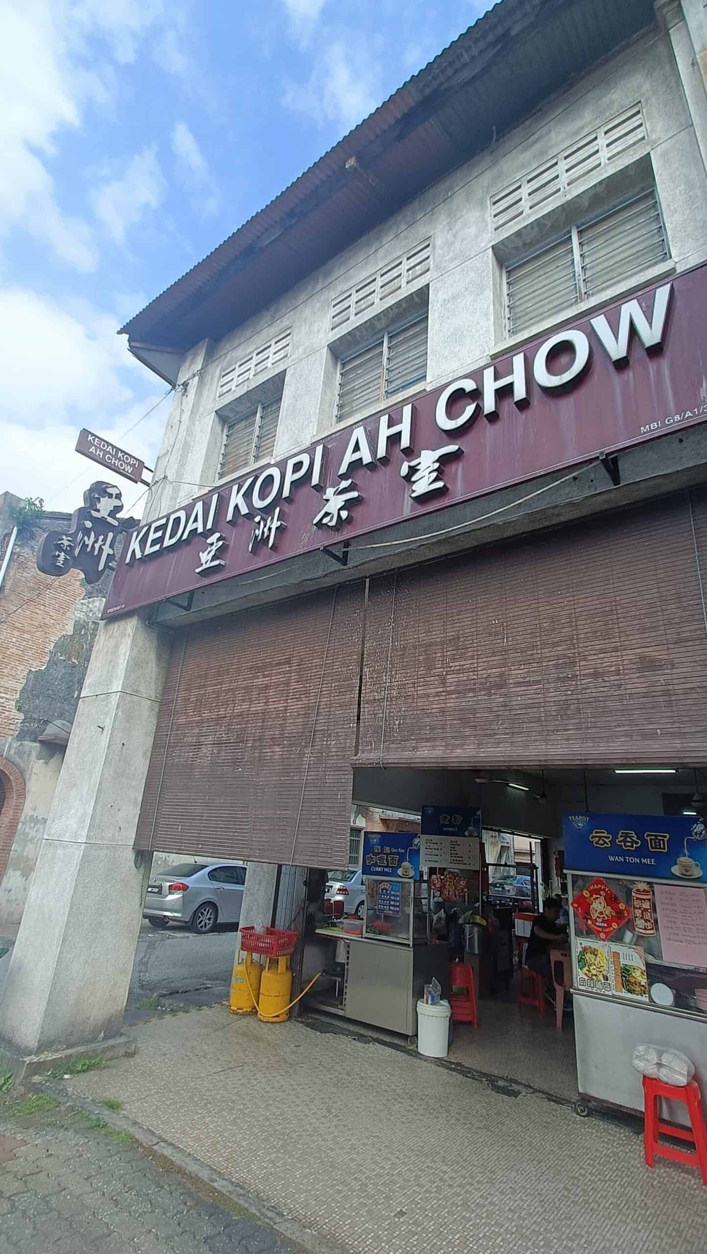 `Exterior of Kedai Kopi Ah Chow in Ipoh with maroon signboard, shutter down, red stools and gas tanks outside`